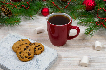 Obraz na płótnie Canvas Red cup with tea, coffee with cookies and marshmallows on a white wooden table against the background of a New Year tree with Christmas decorations. Coziness and New Year concept.