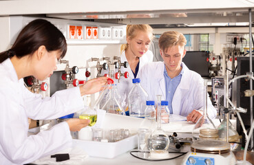 Focused student of Faculty of Chemistry with his female coursemates performing experiments in university laboratory, recording results in workbook