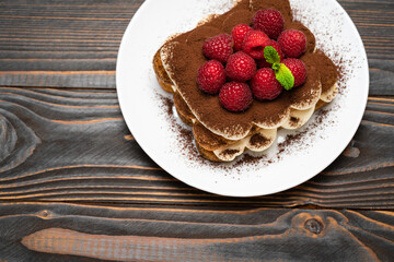 portion of Classic tiramisu dessert with raspberries on ceramic plate on wooden background