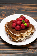 portion of Classic tiramisu dessert with raspberries on ceramic plate on wooden background