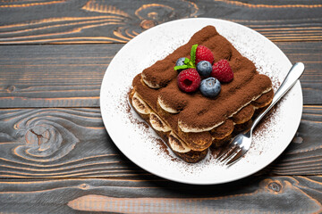 portion of Classic tiramisu dessert with raspberries and blueberries on ceramic plate on wooden background