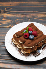 portion of Classic tiramisu dessert with raspberries and blueberries on ceramic plate on wooden background