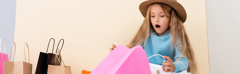 shocked fashionable blonde girl in brown hat, blue sweater looking inside colorful shopping bag near beige wall, panoramic shot