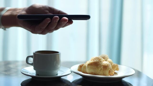 Closeup side view of woman holding modern smartphone and taking trandy flatlay pictures of her morning breakfast coffee and tasty croissants buns to share photos on social media resources