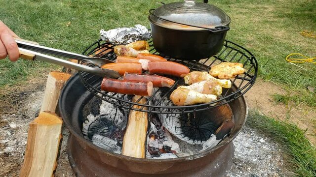 Sausage being turn over or flipped using tongs while being cooked over fire pit on grill alongside chicken drumstick and soup in cast iron pot.
