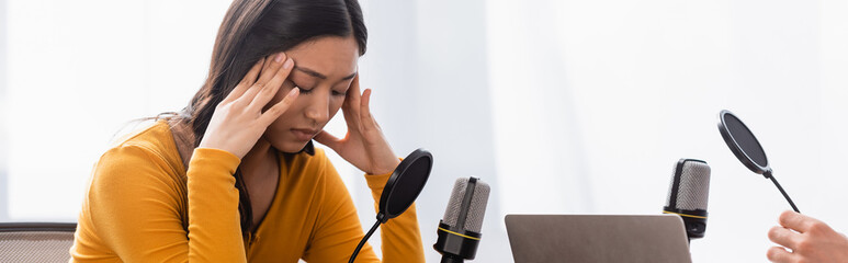 radio host near upset asian woman with closed eyes touching head during interview, horizontal...
