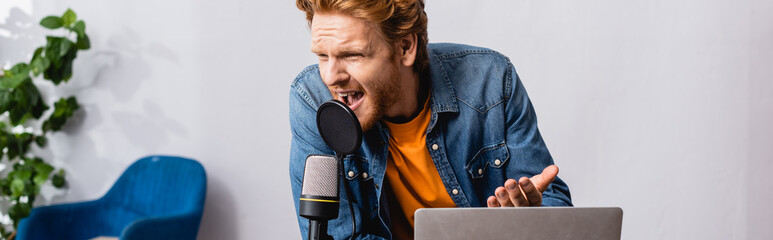 horizontal concept of irritated redhead announcer gesturing while shouting in microphone near laptop