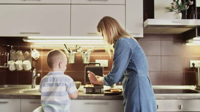 Young mother pulls out cooked waffles from waffle maker and gives plate with it to her son