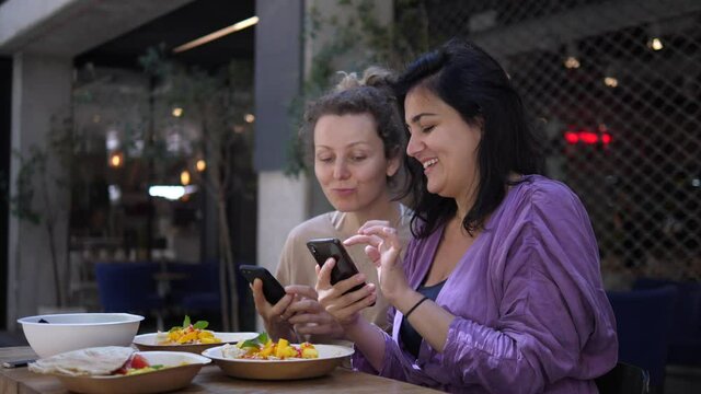 Two cheerful girlfriends showing each other something funny in their phones during lunch. Enjoying delicious healthy food and quality time spent with friends. 