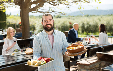 Hospitable young owner of cosy country restaurant in open air meeting guests with serving tray in...
