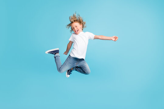 Photo of little child jump raise legs stick-out tongue wear white shirt jeans sneakers isolated blue color background