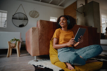 Attractive african female at home sitting on floor using digital tablet and headphones looking away