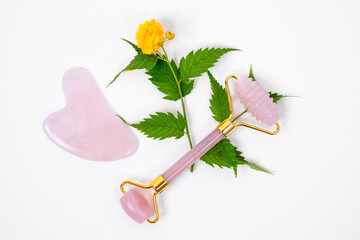 Close up two facial massagers made of natural pink stone, one of flat intricate shape Gua sha, other roller, lying down on white background next to a yellow fresh flower. Copy space.