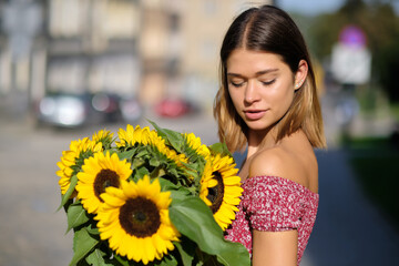 Naklejka premium Young happy beautiful girl is walking in dress with a bouquet of sunflowers on a city street. Autumn concept