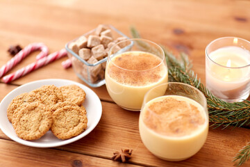 christmas and seasonal drinks concept - glasses of eggnog with oatmeal cookies, candy canes, sugar, fir tree branches and candle burning on wooden background