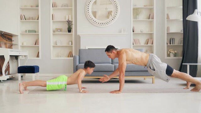Active family doing morning exercise in living room for healthy lifestyle. Dad teaching cute small child boy how to do pushups. Fit sporty father and son play sports at home.