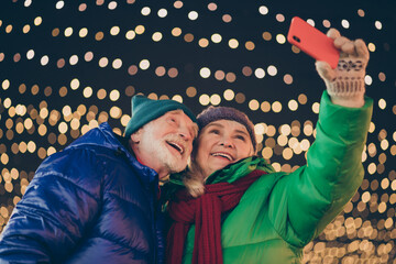 Photo of two people retired friends man woman gathering hold telephone take shoot selfie happy relationship wear coat red scarf headwear x-mas night street illumination fair outdoors outside