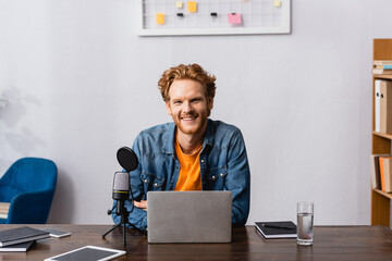 excited redhead announcer looking at camera near gadgets, microphone and notebooks