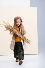 fashionable blonde girl in autumn outfit walking with wheat spikes on beige and white background