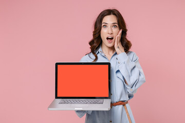 Amazed young brunette woman 20s in casual blue shirt dress hold laptop computer with blank empty screen mock up copy space put hand on cheek isolated on pastel pink colour background studio portrait.