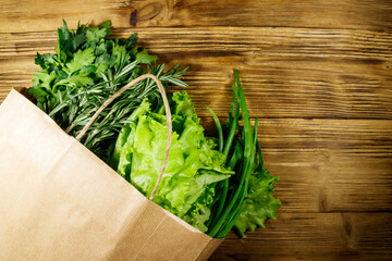 Paper bag with green onion, rosemary, lettuce leaves and parsley on wooden table. Top view. Healthy food and grocery shopping concept