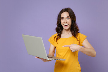 Cheerful young brunette woman 20s wearing basic yellow t-shirt hold in hands pointing index finger on laptop pc computer looking camera isolated on pastel violet colour background, studio portrait.