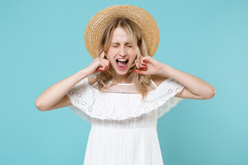 Crazy frustrated young blonde woman 20s in white summer dress hat standing covering ears with fingers keeping eyes closed screaming isolated on blue turquoise colour background studio portrait.