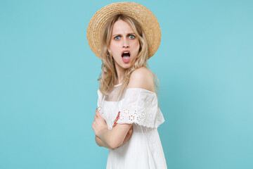 Side view of shocked worried irritated young blonde woman 20s in white summer dress hat standing holding hands crossed looking camera isolated on blue turquoise colour background studio portrait.