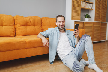 Smiling attractive young bearded man in casual white t-shirt blue shirt using mobile cell phone typing sms message sitting at floor near couch resting relaxing spending time in living room at home.