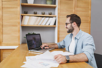 Handsome smiling attractive successful young bearded business man 20s wearing blue shirt glasses sitting at desk with papers document working on laptop pc computer at home or office.