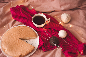 Fototapeta na wymiar Cup of black tea with pink knitted cloth, tasty baked pie and yarning in bed with glowing Christmas lights. Selective focus. WInter holiday season. Good morning.