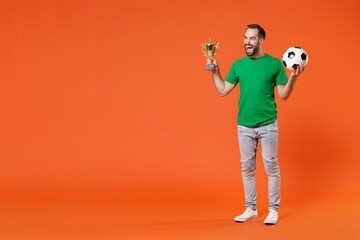 Full length portrait joyful young man football fan in green t-shirt cheer up support favorite team with soccer ball hold golden cup isolated on orange background studio. People sport leisure concept.