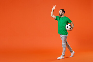 Fototapeta na wymiar Full length portrait of funny man football fan in green t-shirt cheer up support favorite team with soccer ball waving greeting with hand isolated on orange background. People sport leisure concept.