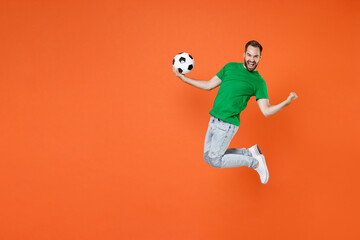 Fototapeta na wymiar Full length portrait happy man football fan in green t-shirt cheer up support favorite team with soccer ball jumping doing winner gesture isolated on orange background. People sport leisure concept.