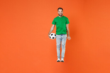 Fototapeta na wymiar Full length portrait of smiling man football fan in green t-shirt cheer up support favorite team with soccer ball jumping isolated on orange background studio. People sport leisure lifestyle concept.
