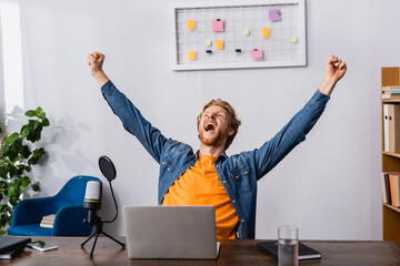excited radio host showing triumph gesture while screaming near microphone and laptop