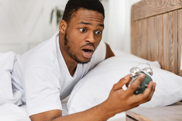 Overslept African Man Holding Alarm-Clock Waking Up In Bed Indoor