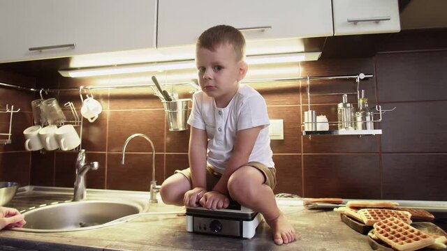 Little boy sits on waffle maker at kitchen
