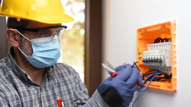 Electrician at work on an electrical panel protected by helmet, safety goggles and gloves; wear the surgical mask to prevent the spread of Coronavirus. Construction industry. Covid-19 Pandemic Prevent