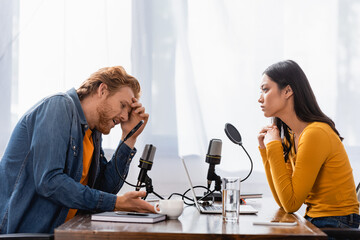 young asian radio host looking at tense man touching head during interview in studio
