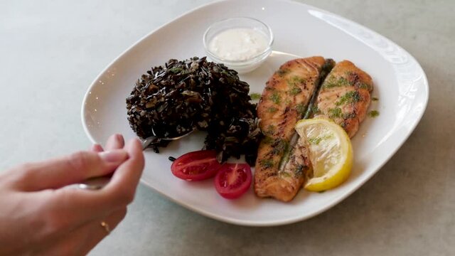 dish with fish and black rice on a white plate. woman eating fish dinner in a restaurant