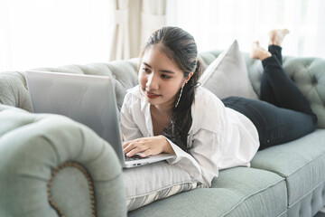 Fototapeta premium Happy casual beautiful Asian woman working on a laptop sitting on the couch in the house.