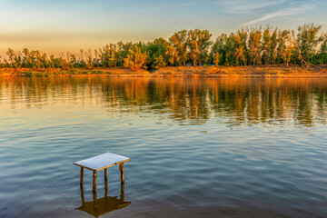 Picturesque summer landscape with riverbank at sunset. Calm plain river, empty table in water....