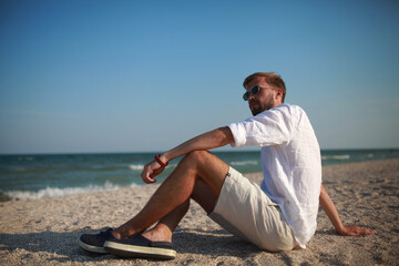 Fototapeta na wymiar Young man sits on beach against sea and sky background.
