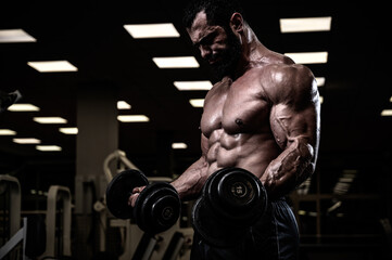 strong young bearded male lifting heavy weight dumbbells training his biceps muscles in dark gym during hard workout