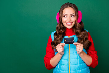 Photo of cute charming young lady hold credit card interested look copyspace presenting good variant smiling wear pink ear warmers blue vest red sweater isolated green color background