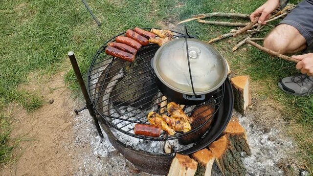 High angle view of fish soup being cooked in cast iron pot over fire pit alongside sausage and chicken drumsticks while more wood sticks being added into camp fire.