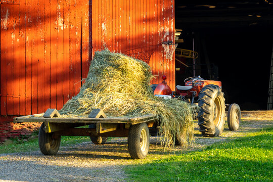 Vintage tractor with a wagon attached loaded with a round bale of hay.  Red bar and basketball hoop visible.  Room for text and great for a background of a farm yard / house.  Country picture.