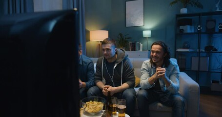 Male football fans sitting on couch in front of TV screen and cheering for favorite team with beer and snacks at night. Guys celebrating win in match and hugs while spending time together in evening.