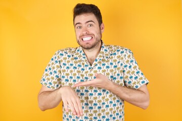 Young man wearing hawaiian shirt over yellow isolated background In hurry pointing to watch time, impatience, upset and angry for deadline delay.